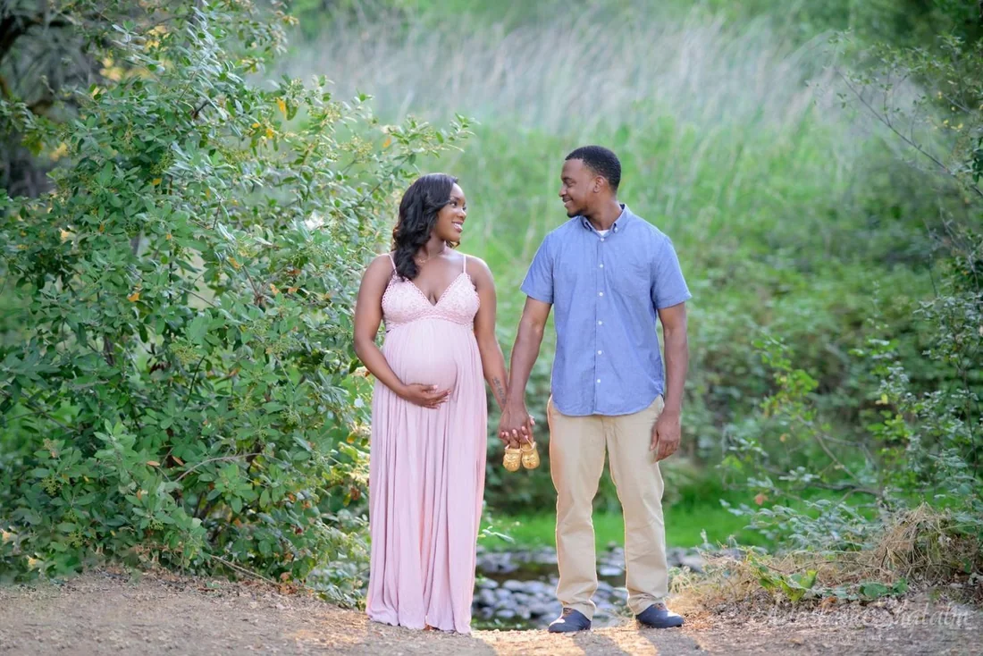 Expectant parents holding hands beside a stream, with a little pair of baby shoes between them.