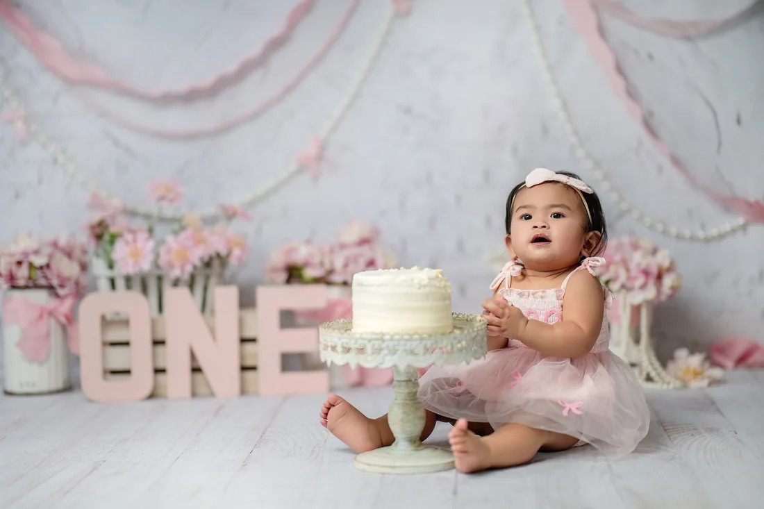 Baby in a pink dress beside a white first birthday cake