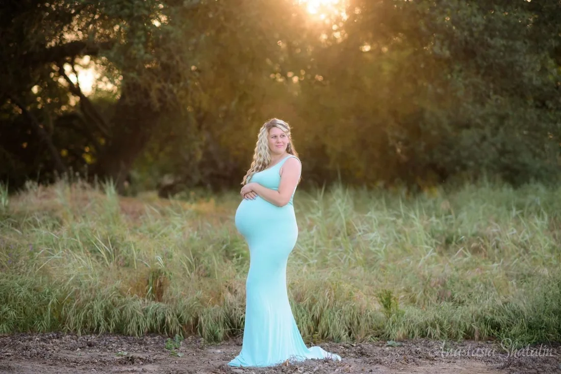 An outdoor maternity portrait in a flowing turquoise dress, with warm evening light falling through the trees.