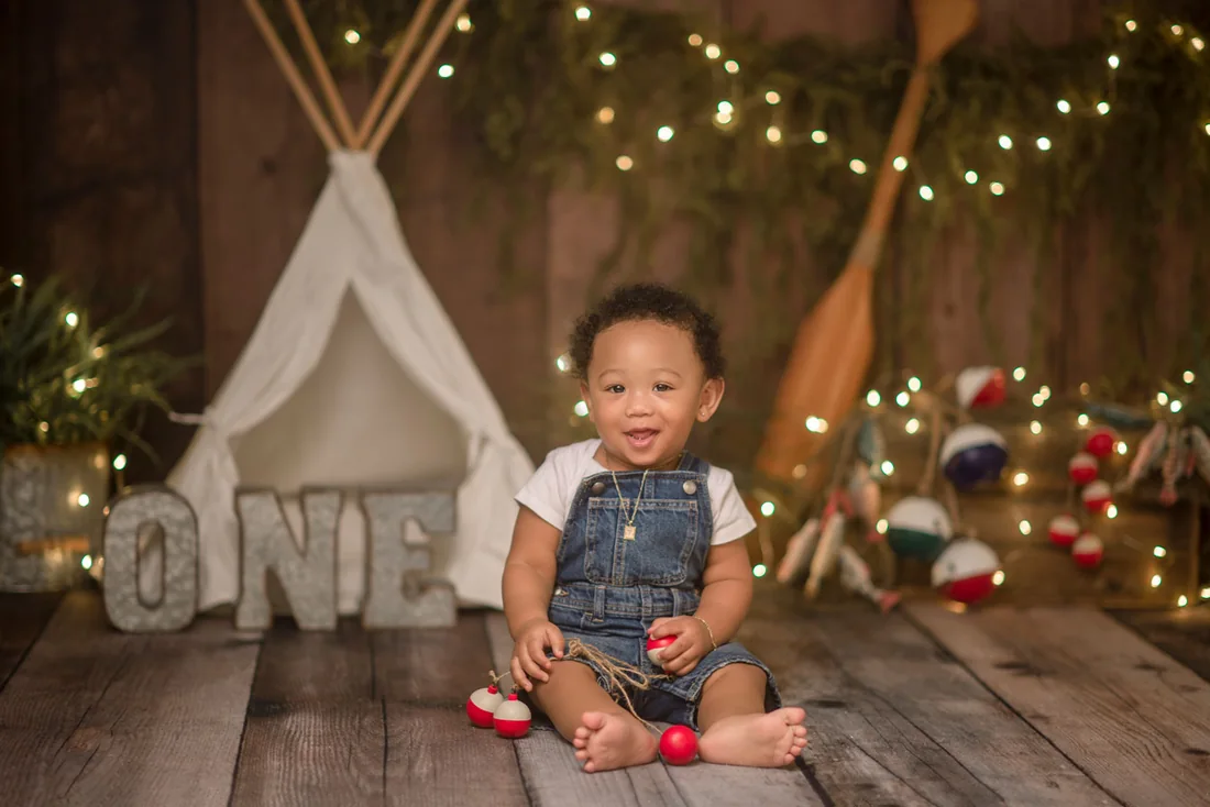 Smiling birthday boy in denim overalls beside a tent and ONE letters