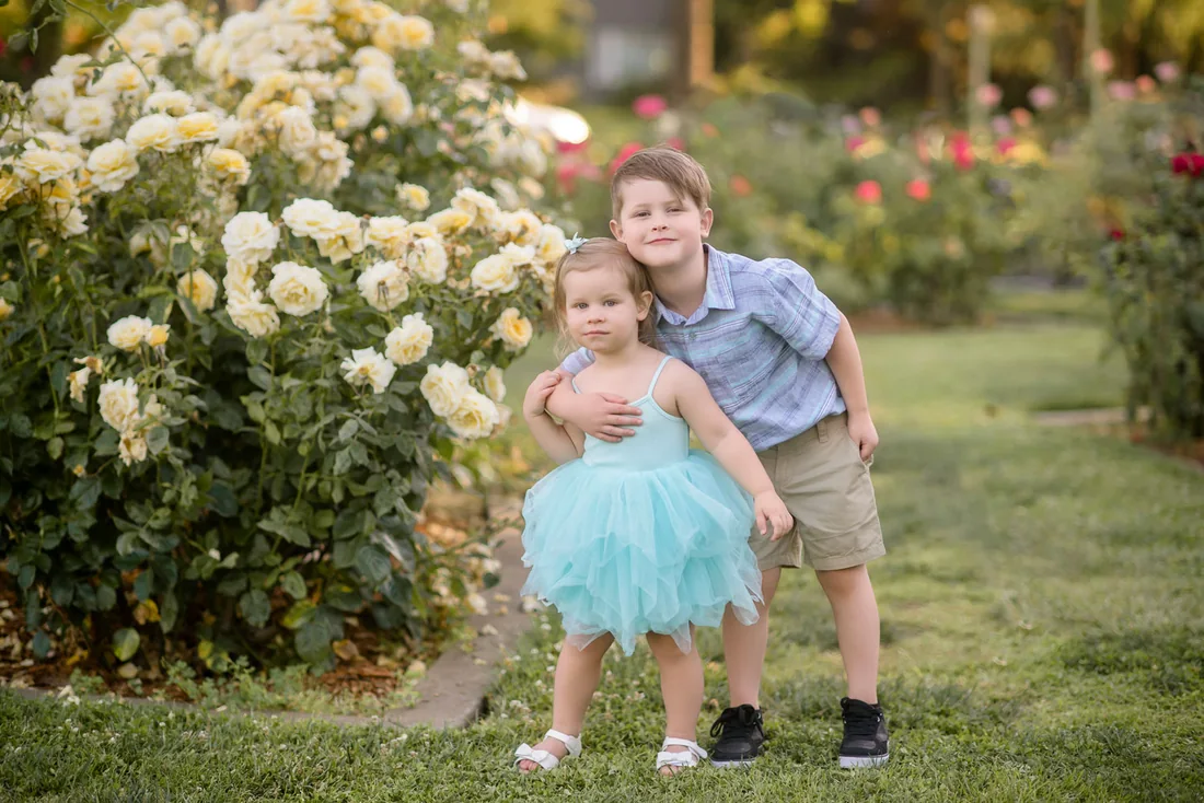 Two children standing beside a white rose bush